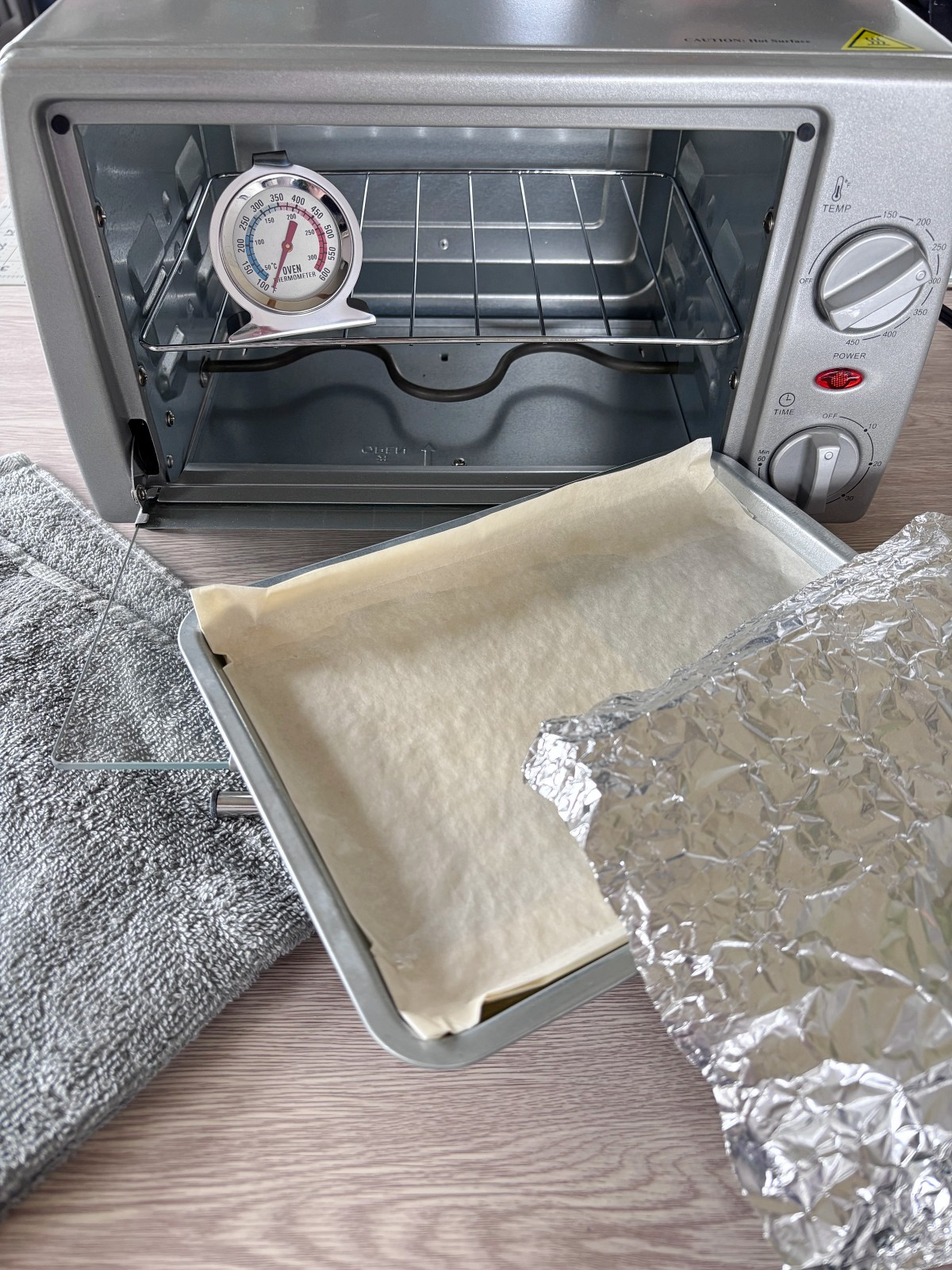 Toaster oven with oven thermometer, foil, parchment paper, and a towel on a table.